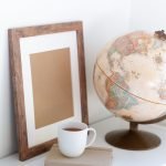 Vintage globe near blank frame and cup of tea placed on book in office